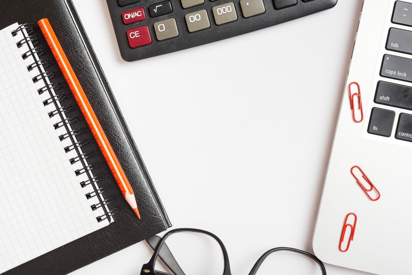 Ein Schreibtisch auf dem ein Notizblock, ein Taschenrechner und ein Laptop liegen.